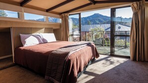 A bedroom with a large bed, a view of mountains through a sliding glass door, and a balcony with a view of a house and mountains.
