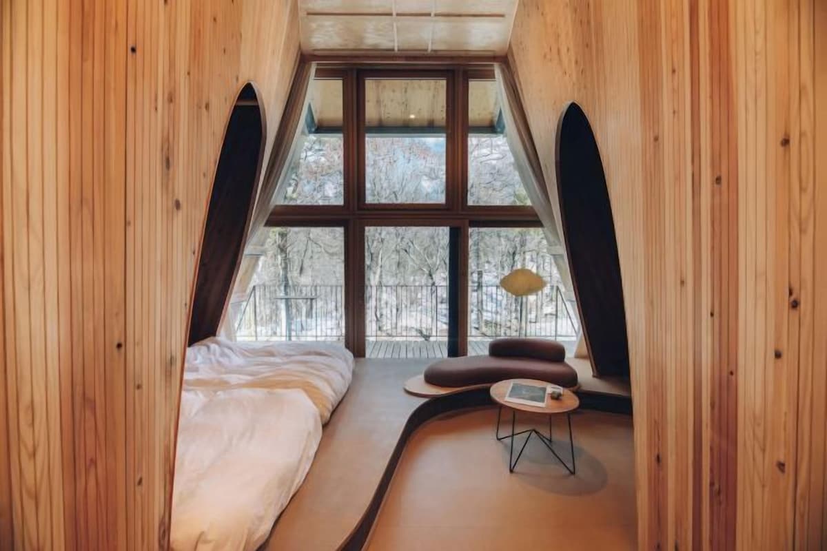 A cozy bedroom with a bed, a small table, and a window with a view of trees.
