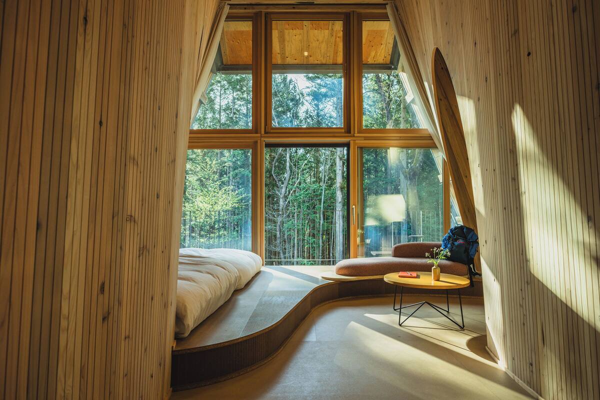 A bedroom with a curved bed, a small round table with a plant, and a view of a forest through large windows.