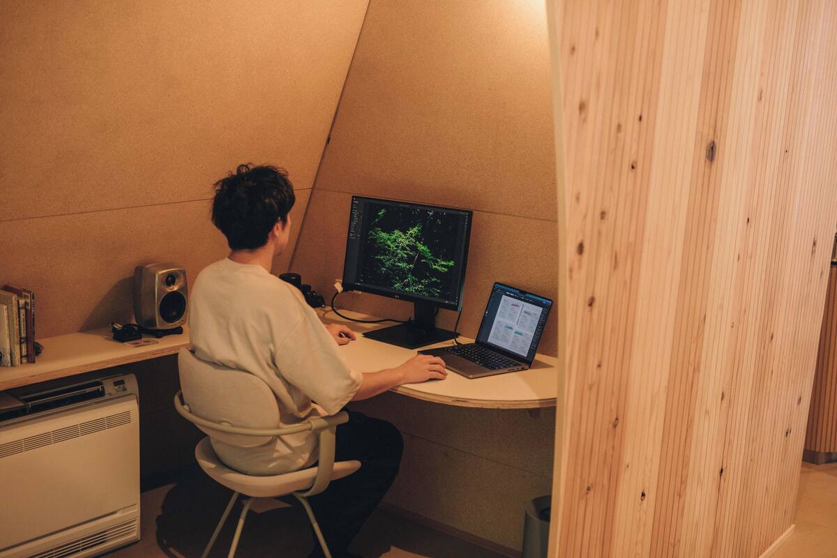 A person seated at a desk with a computer, a speaker, and a bookshelf in a room with wooden paneling.