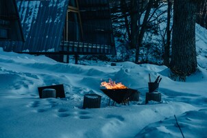 Un espacio al aire libre cubierto de nieve, con un hoyo de fuego, leña y una estructura metálica al fondo.