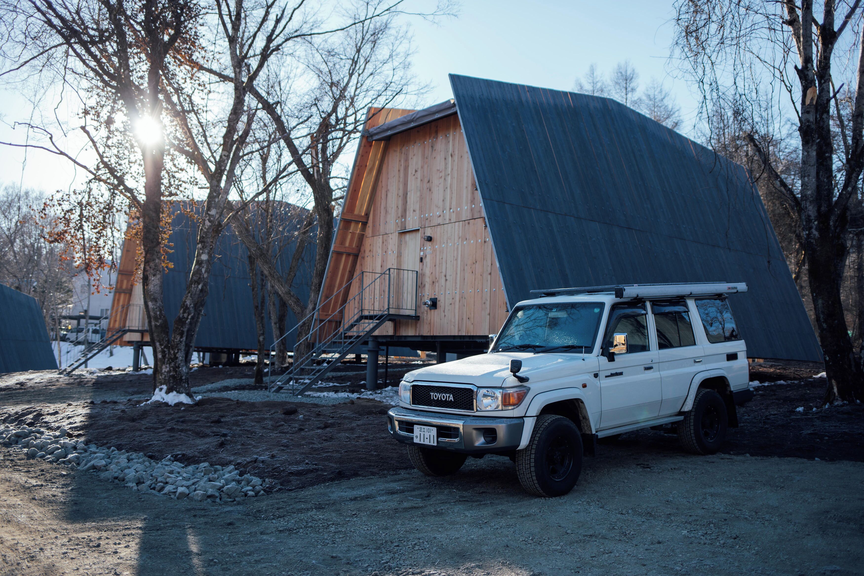 A white SUV parked in front of a large wooden structure with a sloped roof, surrounded by trees and a dirt ground.