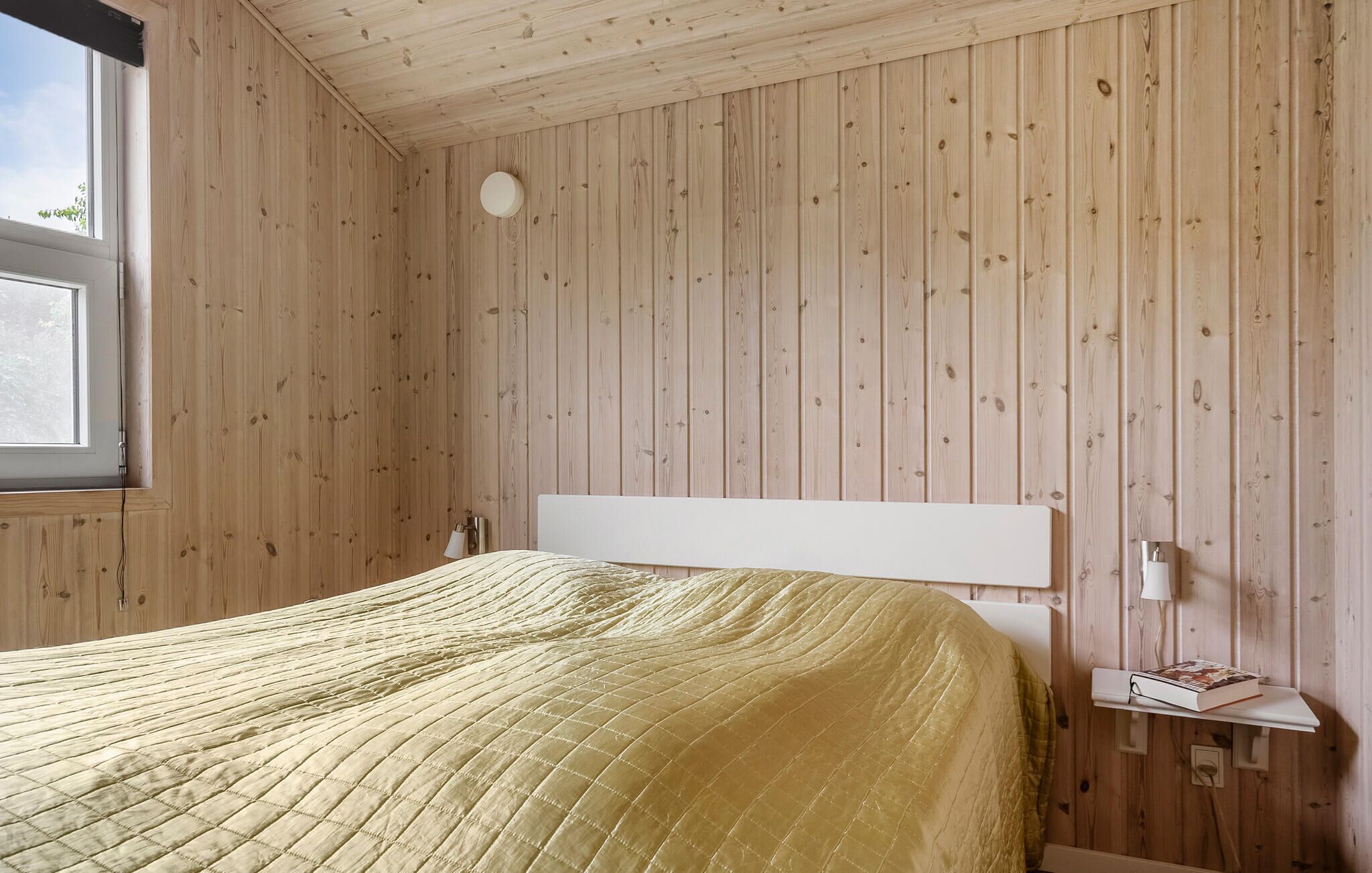 Een kamer met een bed, houten wanden, een raam en een nachtkastje met een boek.
