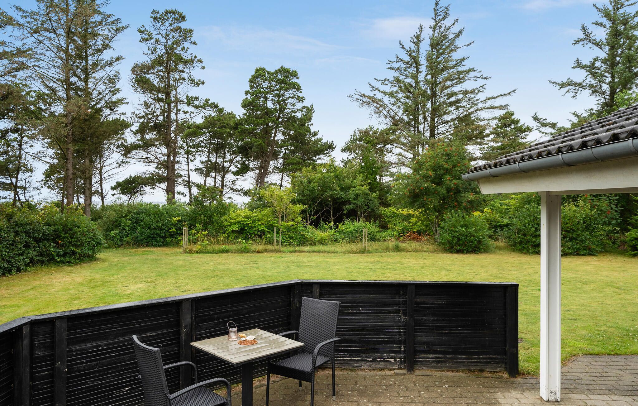 A patio area with a table and chairs, surrounded by a black fence, overlooking a grassy yard with trees and a white structure in the background.