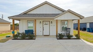 A single-story house with a gabled roof, a covered porch with two chairs, and a small garden area. - 3 bedroom, 2 bath house, sleeps 6+, baby bed W (Hood County)