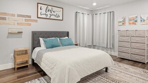 Une chambre à coucher comprenant un lit, une table de chevet, une commode, un ventilateur de plafond et un mur orné d’une citation inspirante.