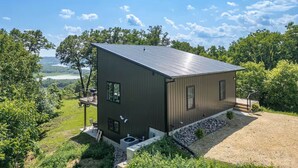 Une maison moderne à deux étages, dotée d’un toit en métal, entourée d’une végétation luxuriante et offrant une vue panoramique sur des collines et un lac.