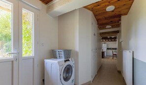A laundry room with a washing machine and a basket on top, a wooden ceiling, and a view into a kitchen area.