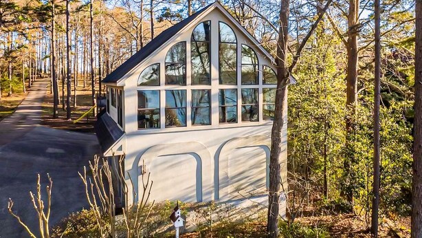 A small, white, single-story building with large windows, surrounded by trees and a paved path. - One-bedroom treehouse loft with lake views, shared dock access, and full kitchen on Lake Hamilton. (Garland County)