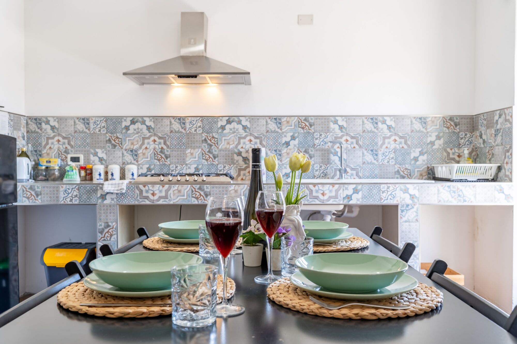 Una mesa de comedor dispuesta con platos verdes, vasos y una botella de vino tinto, con un salpicadero decorado y una campana extractora moderna al fondo.