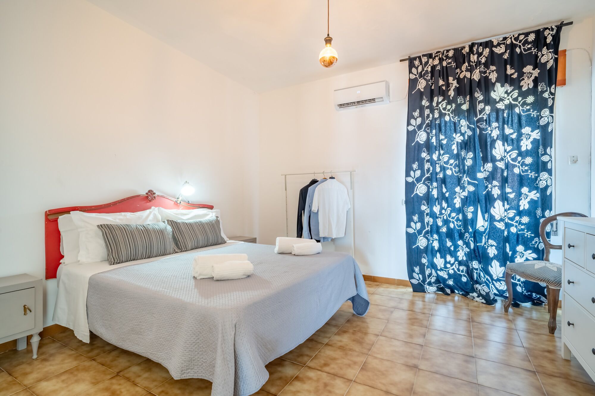 A bedroom with a bed, bedside tables, a hanging light, a wardrobe, and a window with a floral curtain.
