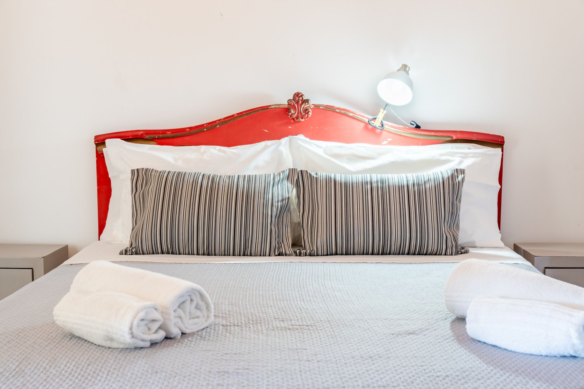 A neatly made bed with striped pillows and a white blanket, a red headboard, and a bedside lamp.