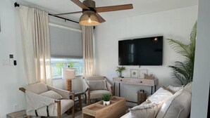 Ein modernes Wohnzimmer mit einer Couch, zwei Sesseln, einem Couchtisch, einem Fernseher und einem Deckenventilator.