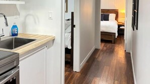 A narrow hallway with a kitchen on one side and a bedroom on the other. The kitchen has a sink, cabinets, and a dishwasher. The bedroom has a bed, a nightstand, and a window with curtains. - Lakefront Getaway Sparrow Lake Muskoka Beach House (Gravenhurst)