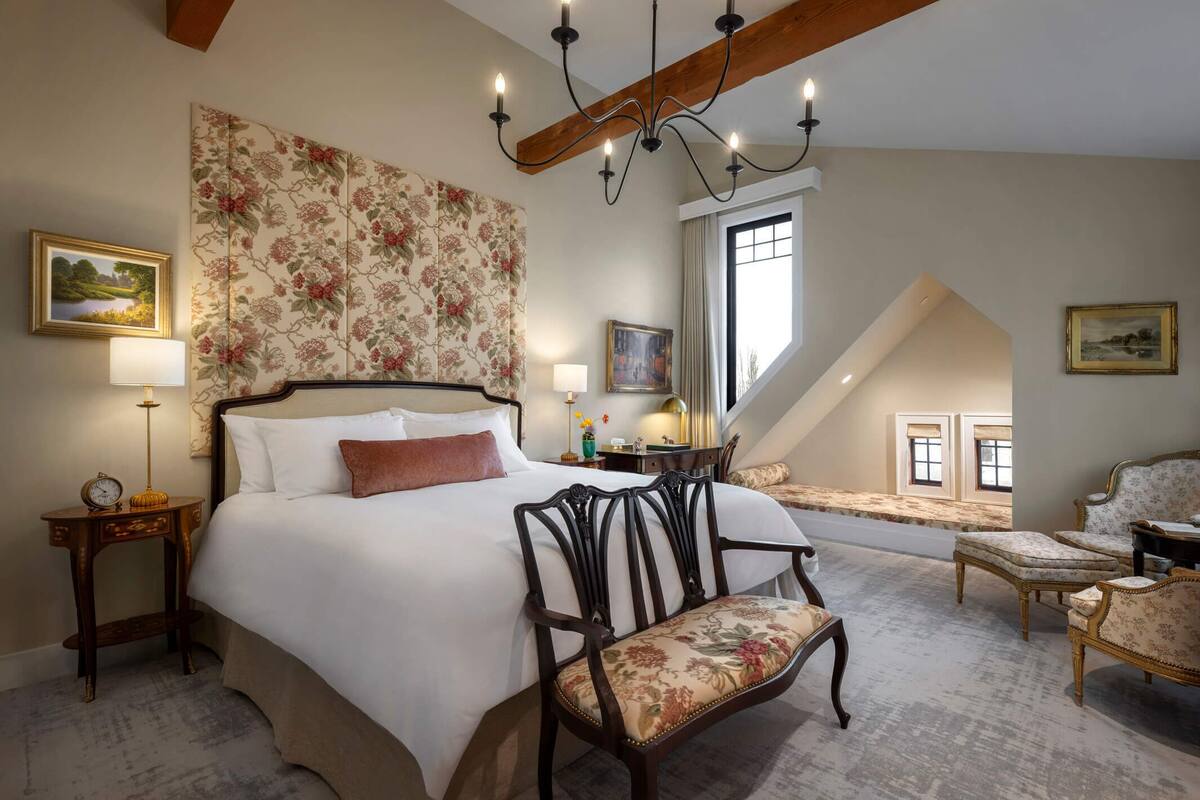 A bedroom with a large bed, a wooden chair, a chandelier, and a view of the outside.