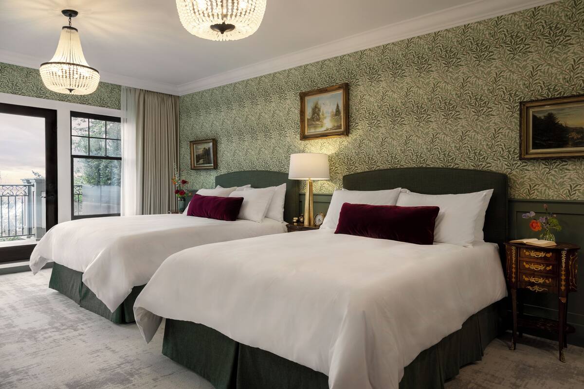 Une chambre d’hôtel avec deux lits, un lustre, une fenêtre donnant sur un beau paysage et un papier peint à motifs.