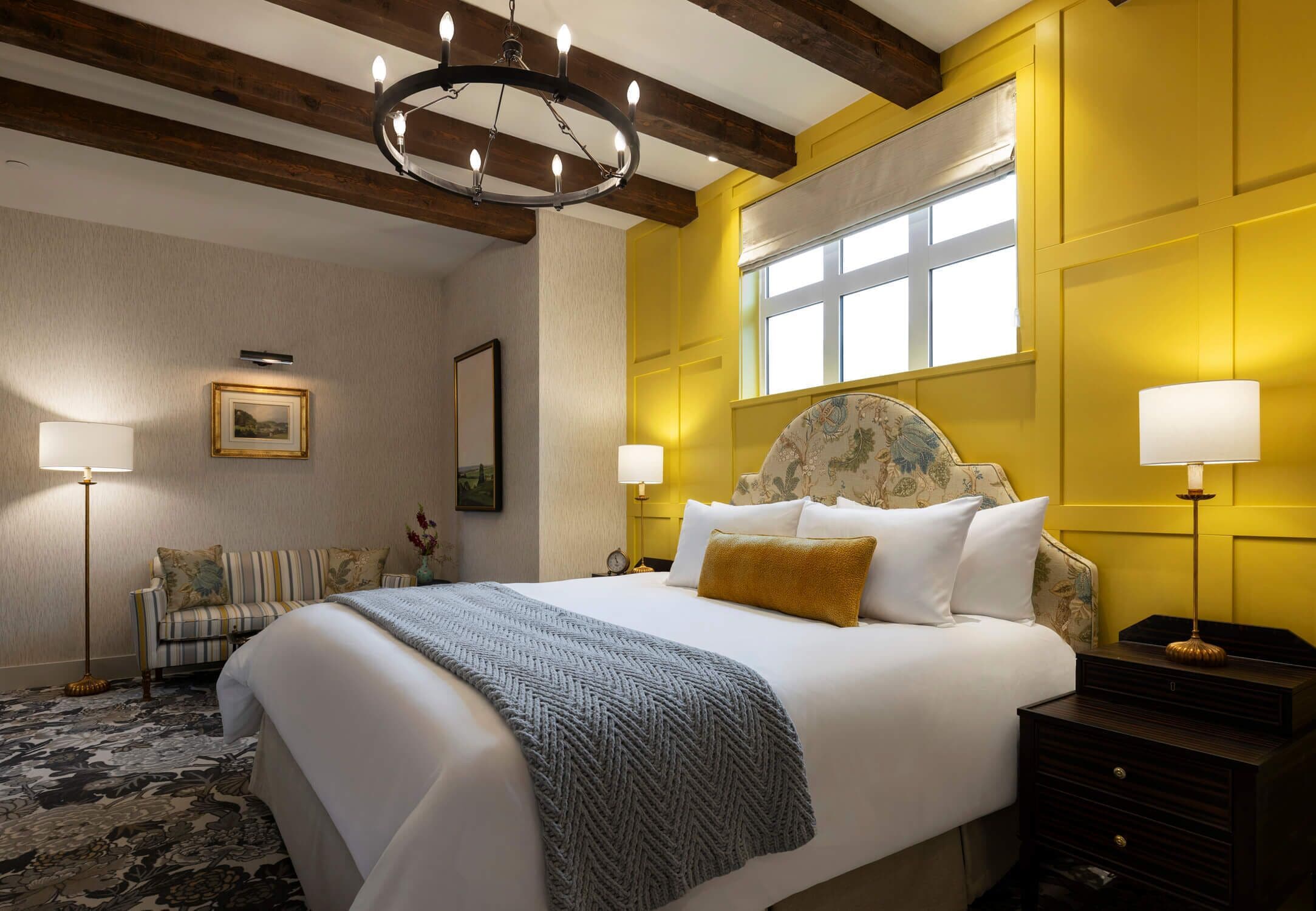 Een hotelkamer met een groot bed, een kroonluchter, een raam met jaloezieën, een nachtkastje en een vloerlamp.