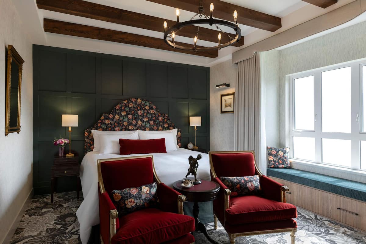 Une chambre d’hôtel avec un grand lit, deux fauteuils rouges, un lustre, une fenêtre avec vue et une tête de lit décorative.
