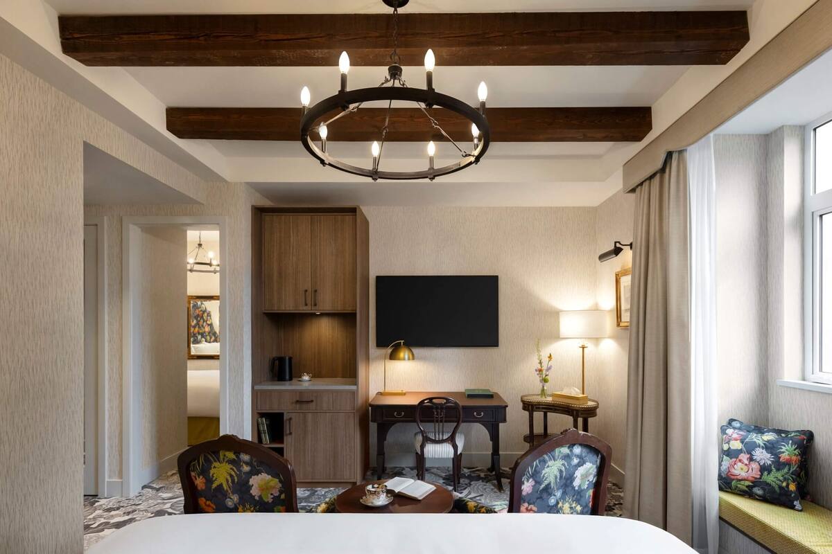 Une chambre d’hôtel comprenant un lit, un bureau, une chaise, une petite table, une bibliothèque, un téléviseur à écran plat, une lampe et une fenêtre avec des rideaux.