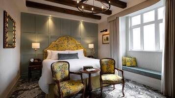 Habitación de hotel con una cama grande, dos sillas, una mesita, una ventana con cortinas y un candelabro.