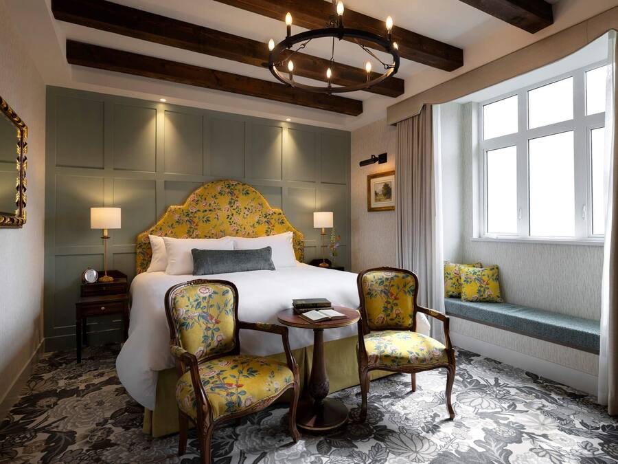 A hotel room with a large bed, two chairs, a small table, a window with curtains, and a chandelier.