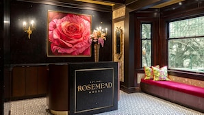 Ein Empfangsbereich mit einer schwarzen Rezeption, auf der der Name „ROSEMEAD HOUSE“ steht, und einem großen gerahmten Bild einer Rose. Ein Fenster bietet Blick auf Bäume, und eine Bank ist mit bunten Kissen ausgestattet.