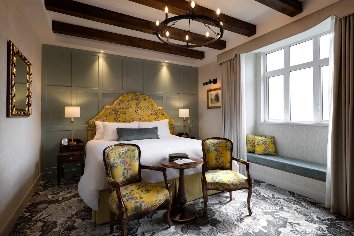 Une chambre d’hôtel avec un grand lit, deux fauteuils, une petite table, une fenêtre avec des rideaux et un lustre.