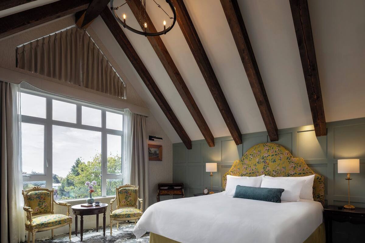 Une chambre avec un grand lit, deux fauteuils, un lustre et une vue sur l’extérieur grâce à une grande fenêtre.