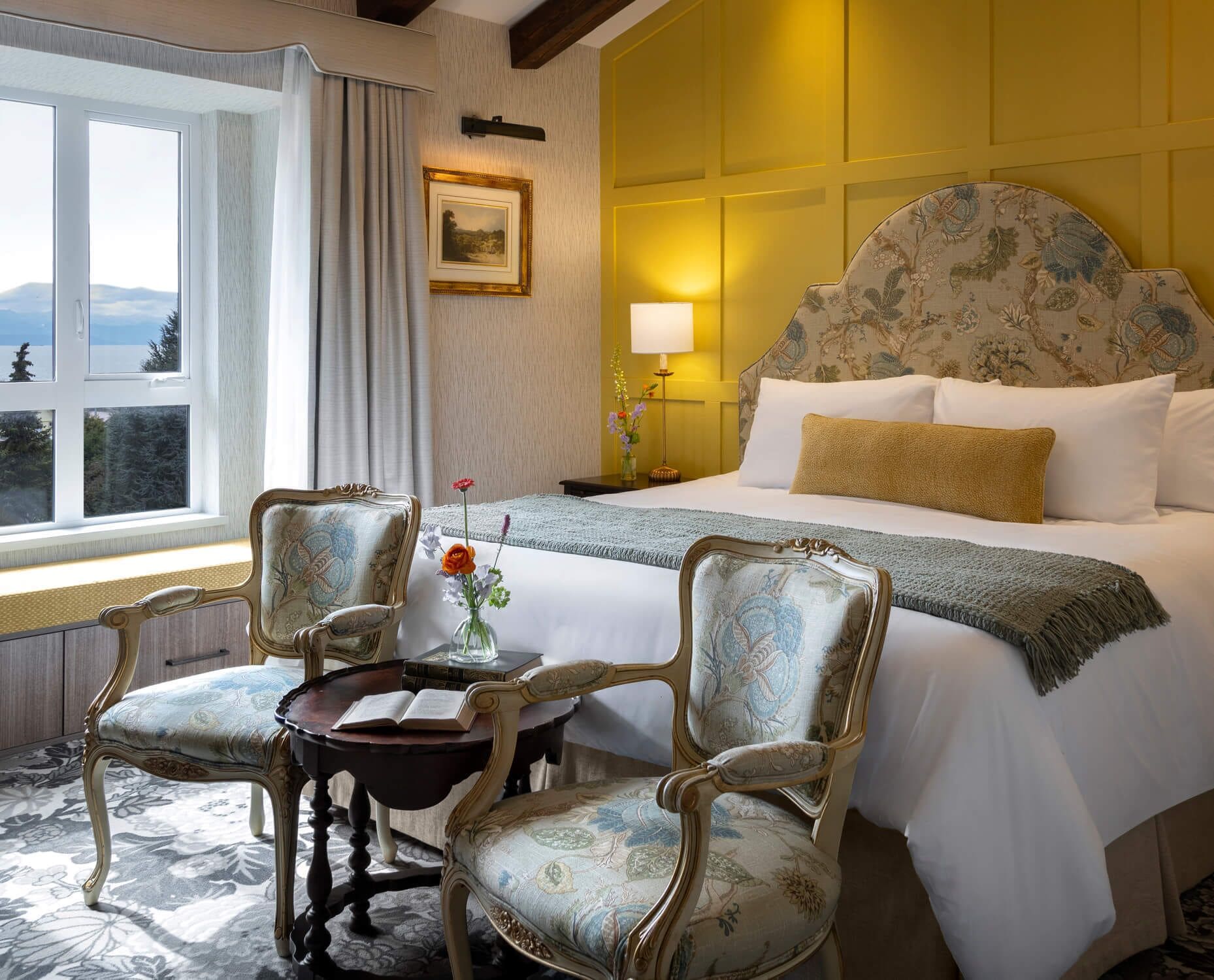 Ein Hotelzimmer mit einem großen Bett, zwei Polstersesseln, einem kleinen Tisch mit Blumenvase, einem gerahmten Bild an der Wand und Bergblick durch das Fenster.