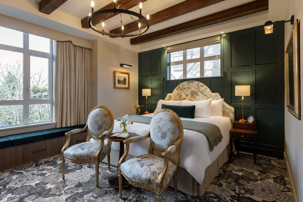 A hotel room with a large bed, two upholstered chairs, a small table, a chandelier, and a view through a window.