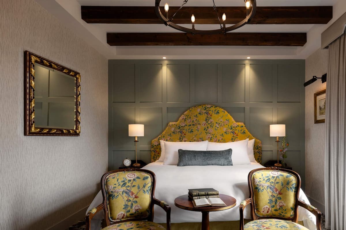A hotel room with a large bed, two chairs, a small table, a mirror, and a window with curtains.
