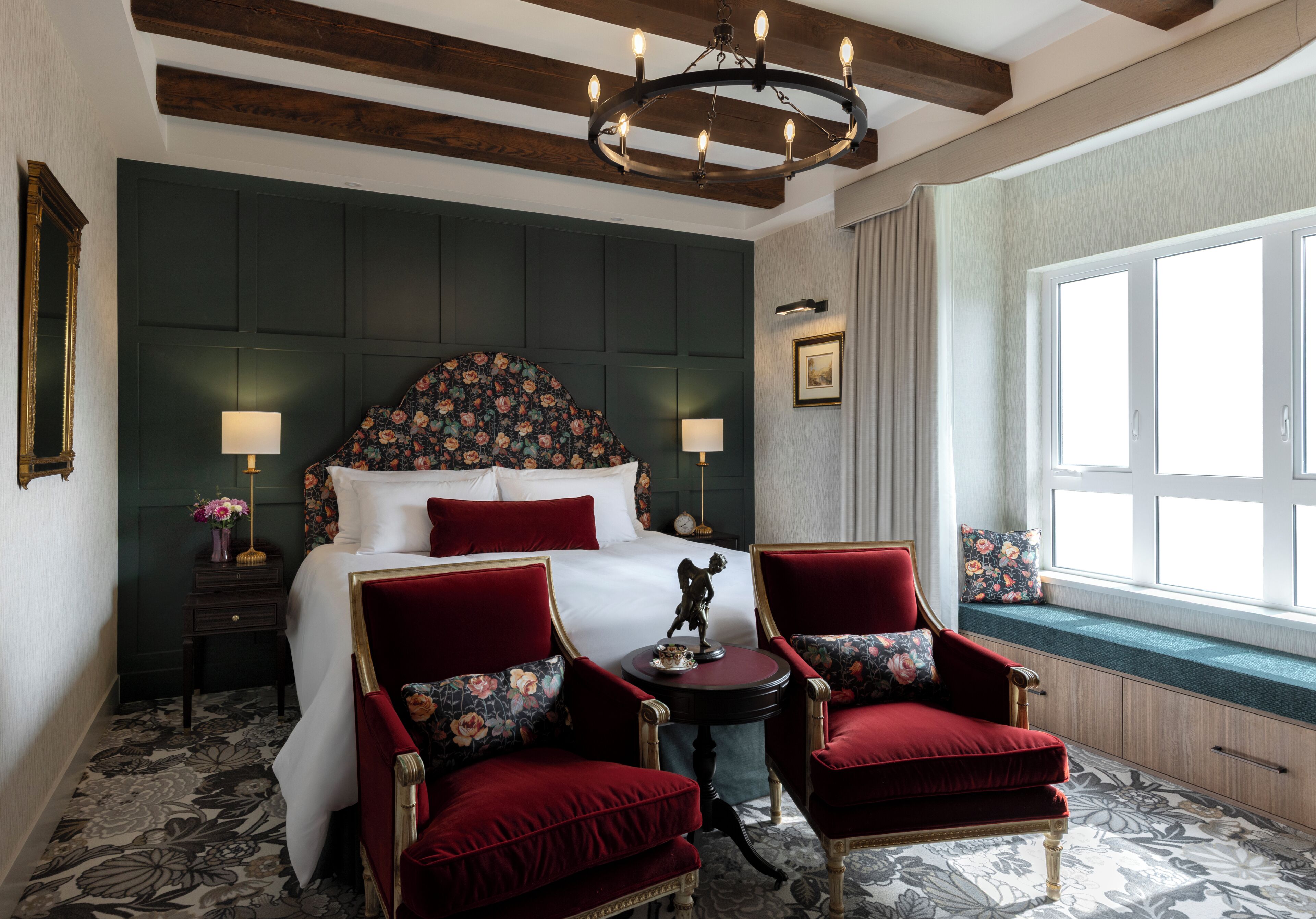 A hotel room with a large bed, two red upholstered chairs, a chandelier, and a window with a view.