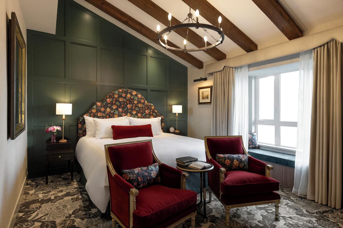 Une chambre d’hôtel avec un grand lit, deux fauteuils rouges, une petite table, un lustre et une fenêtre avec des rideaux.