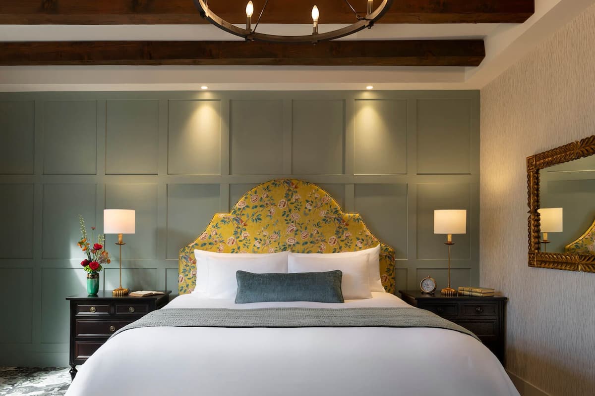 Une chambre d’hôtel avec un grand lit, une tête de lit à motifs floraux, des lampes de chevet, un miroir et une table de chevet avec une horloge.