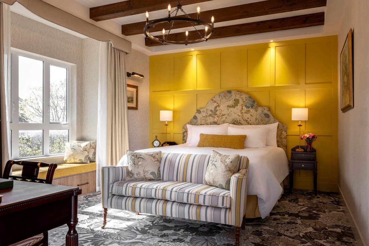 A hotel room with a bed, a sofa, a nightstand, a lamp, a window, and a chandelier.
