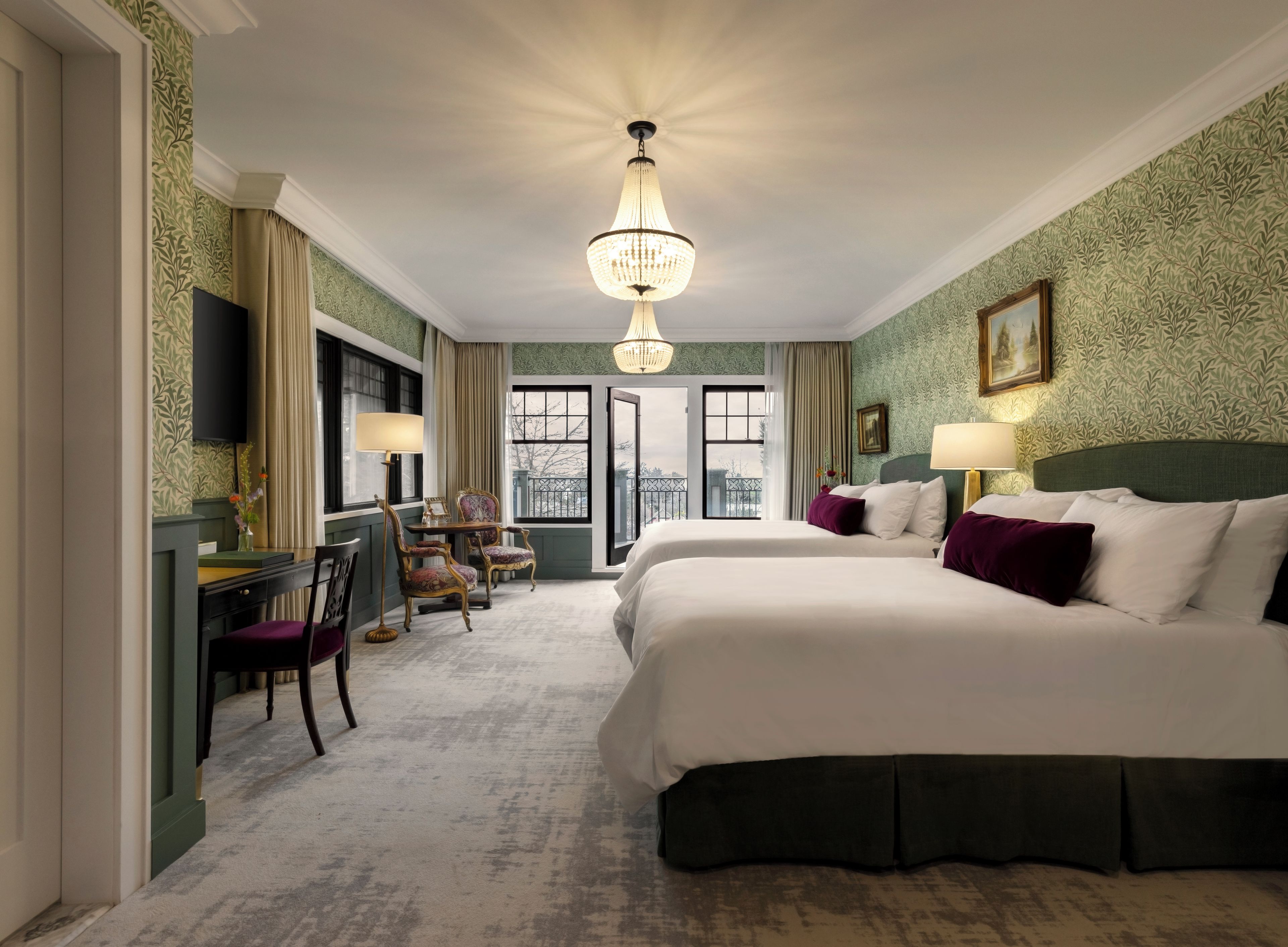 Ein Hotelzimmer mit zwei Betten, einem Schreibtisch, Stühlen, einer Chandelier und Blick durch die Tür.