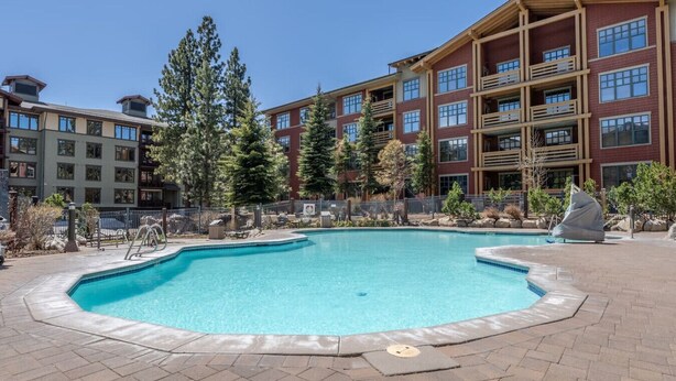 A pool area with a clear blue pool, surrounded by a brick patio and a multi-story building with balconies.