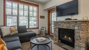 A living room with a stone fireplace, a large flat-screen TV mounted above it, a gray sofa with cushions, and a round coffee table with a book.