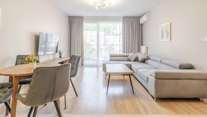 Una sala de estar moderna con un sofá, una mesa de comedor con sillas, un televisor y un amplio ventanal con cortinas.