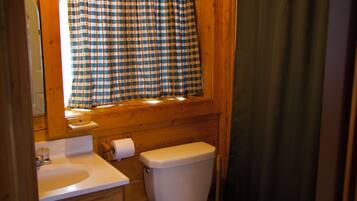A bathroom with a wooden interior, a window with a checkered curtain, a toilet, and a sink with a roll of toilet paper.