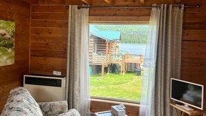 A wooden cabin room with a large window, a sofa, a TV, and an air conditioning unit. - McNeil River Cabin, Homer Floatplane Lodge (Homer)