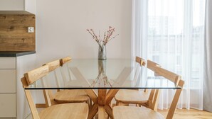 A modern dining area with a glass table surrounded by wooden chairs, a vase with flowers, and a view of the city through sheer curtains.