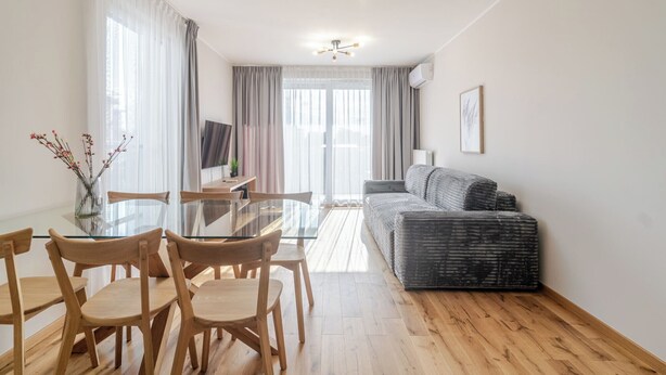 A modern living room with a glass dining table, wooden chairs, a grey sofa, and a TV on the wall.