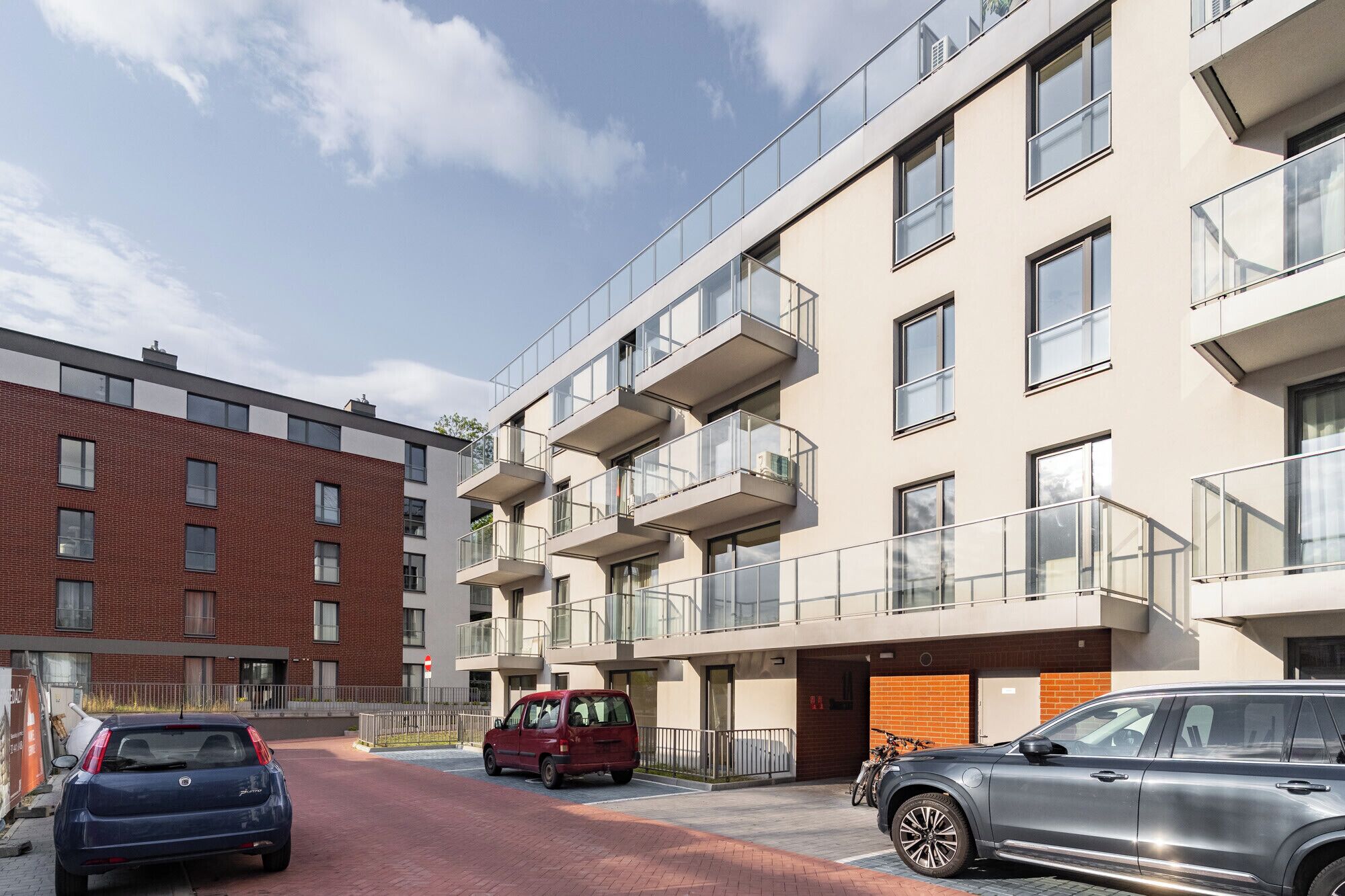 Een modern appartementencomplex met meerdere balkons, een parkeerplaats en een rode auto ervoor geparkeerd.
