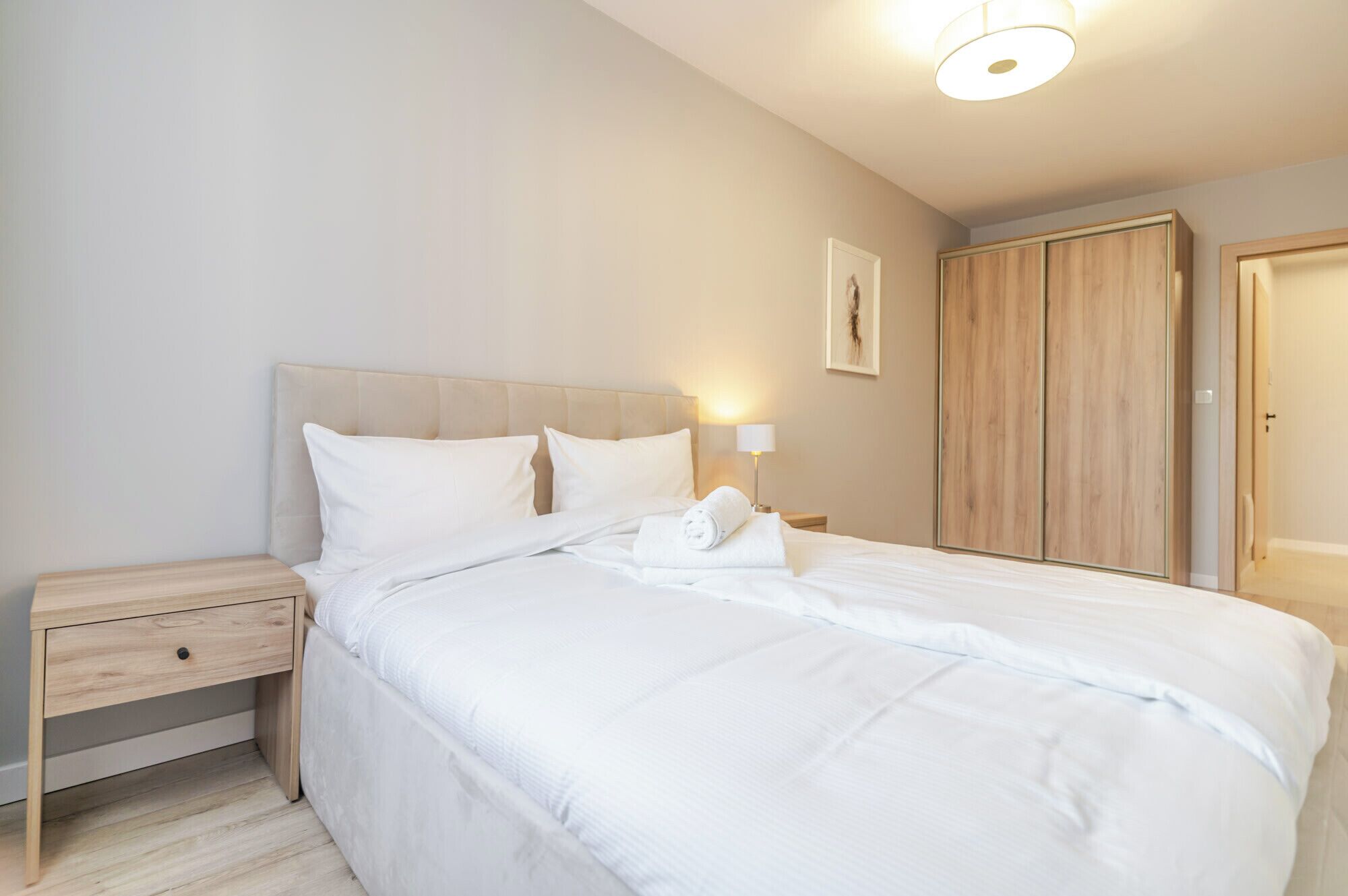 A neatly made bed with white linens, a bedside table with a lamp, and a wooden wardrobe.