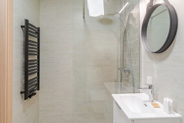 A modern bathroom with a black towel rack, a glass shower enclosure, a white sink with toiletries, and a round mirror.