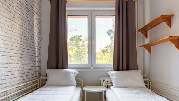 Une chambre avec deux lits simples, une petite table, une fenêtre avec des rideaux, un mur en briques et des étagères en bois.