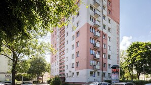 Un immeuble résidentiel à plusieurs étages, doté d’une façade rouge et blanche, de nombreux balcons et d’un espace de stationnement où sont garées plusieurs voitures.