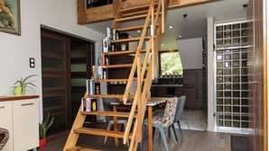 Een houten trap leidt naar een slaapkamer op een entresol, een eethoek met tafel en stoelen, en een keuken met uitzicht.