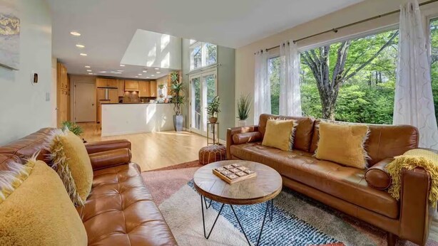 A spacious living room with a leather sofa, a coffee table, and a view of the kitchen and outdoor greenery.